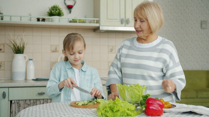 Adorable blond child is helping granny in kitchen making salad doing high-fivepreparing healthy meal for dinner. People, family and apartment concept.